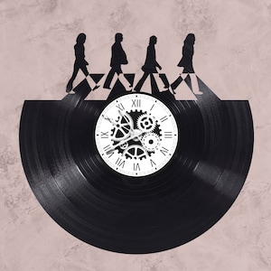 Op de afbeelding: Een zwarte vinyl platenspeler klok met een witte wijzerplaat en tandwielen. De wijzerplaat heeft Romeinse cijfers en de plaat heeft een silhouet van vier mensen die over een zebrapad lopen.
