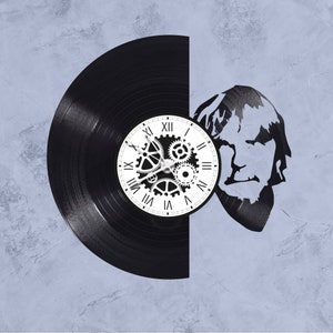 May include: A black vinyl record clock with a white clock face and gears. The clock face has Roman numerals and the gears are in the center of the clock face. The record is cut in half and the other half is a silhouette of a person's head.