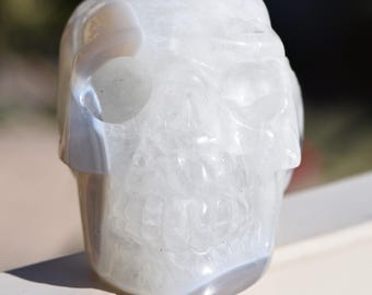 May include: A carved skull sculpture made of translucent white and grey banded agate. The stone features a polished, smooth surface with visible natural mineral layers and inclusions throughout the structure, forming a detailed human skull shape.