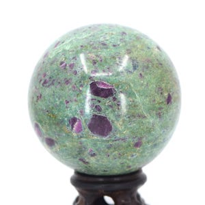 May include: A green and purple gemstone sphere on a wooden stand. The sphere is polished and has a speckled appearance.