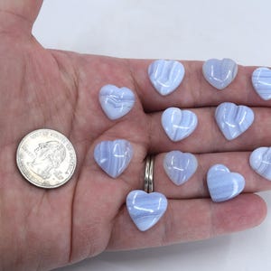 May include: A hand displaying several light blue heart-shaped gemstones. A UK quarter-sized coin is also present for scale. The gemstones have a marbled appearance.