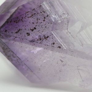 May include: A purple amethyst crystal with a clear, faceted point. The crystal has a natural, rough texture and is filled with small, dark inclusions.