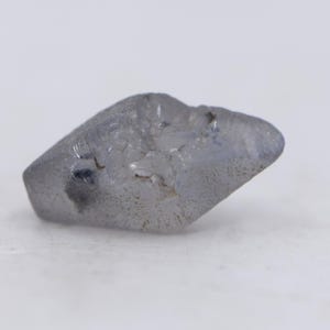 May include: A rough, uncut gemstone with a light blue-gray hue. The stone has a textured surface and irregular shape, with a few darker inclusions. The gem is displayed against a white background.