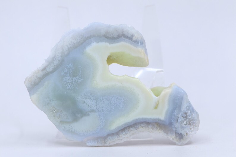 Pu&ograve; includere: Una geode di agata blu chiaro e bianco con una forma unica e curva. La geode ha un esterno naturale e ruvido e un interno liscio e lucido.