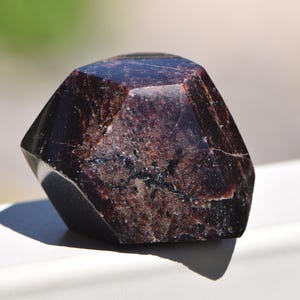 May include: A dark reddish-brown garnet crystal with a faceted, geometric shape. The stone features a polished, reflective surface with visible internal fractures and deep, opaque color variations throughout its solid, angular structure.