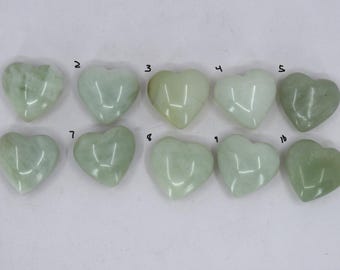 May include: Ten heart-shaped jade stones in varying shades of green are arranged on a white surface. Each stone has a smooth, polished appearance, with subtle variations in colour and veining. The hearts are numbered 1 to 10.
