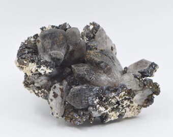 May include: A cluster of smoky quartz crystals embedded in a matrix of gray and black minerals. The crystals are translucent and have a slightly cloudy appearance.