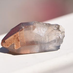 May include: A translucent quartz crystal point featuring a distinct reddish-brown iron oxide inclusion concentrated at one end, transitioning into a clear, glassy termination with natural facets and a jagged, unpolished base structure.