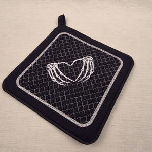Könnte beinhalten: Schwarzer quadratischer Topflappen mit einem weißen, gestickten Design aus Skeletthänden, die ein Herz bilden. Der Topflappen hat einen schwarzen Rand und eine Schlaufe zum Aufhängen. Die Mitte hat ein weißes, genähtes Gittermuster.
