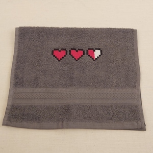 Op de afbeelding: Een grijze handdoek met drie geborduurde rode harten, waarvan er één half gevuld is met wit.