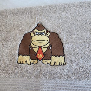 Può includere: Un asciugamano beige con una toppa ricamata centrale del personaggio Donkey Kong. Il personaggio è raffigurato con pelliccia marrone, mani e viso color marrone chiaro e una cravatta rossa con le lettere gialle DK ricamate sul davanti.
