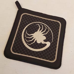 Puede incluir: Un agarrador de olla acolchado negro y marrón con una criatura alienígena bordada en beige dentro de un círculo.