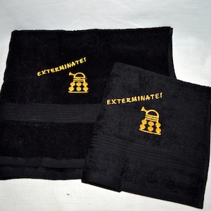 Könnte beinhalten: Zwei schwarze Badetücher mit goldener Stickerei. Die Handtücher zeigen ein Cartoon-Bild eines Daleks aus der Fernsehserie Doctor Who und den Text "Exterminate!"