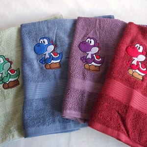 Könnte beinhalten: Vier farbenfrohe Badetücher, jedes mit einem anderen Yoshi-Charakter bestickt. Die Handtücher sind in den Farbtönen Grün, Blau, Lila und Rot gehalten. Die Yoshi-Charaktere sind farblich passend, mit braunen Schuhen und weißen Akzenten.