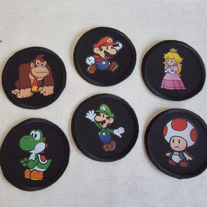 Puede incluir: Seis parches circulares negros con personajes de dibujos animados coloridos. Los personajes incluyen a Mario, Luigi, la Princesa Peach, Yoshi, Toad y Donkey Kong. Los parches son probablemente para ropa o accesorios.