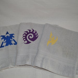 May include: Three white bath towels with embroidered designs. One towel features a blue emblem, another a purple swirl, and the third a yellow flame. The towels are folded and stacked, showcasing the embroidered details.