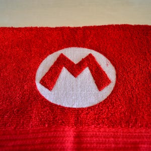 Può includere: Asciugamano da bagno rosso con un logo Mario ricamato in bianco.