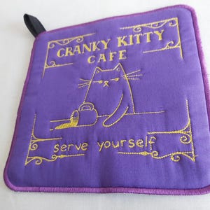 Może przedstawiać: Fioletowy, kwadratowy podkładka pod garnek ze złotym haftem. Tekst brzmi "CRANKY KITTY CAFE" i "serve yourself", z ilustracją kota. Czarna pętla jest przymocowana.