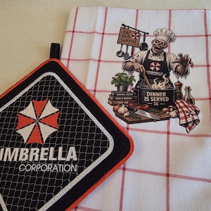 May include: A black square pot holder with an orange border featuring the white Umbrella Corporation logo and text, alongside a white kitchen towel with a red grid pattern and a graphic of a zombie chef with the text Eat Heal Survive.