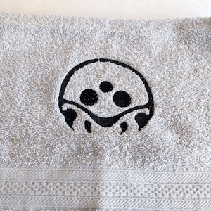 以下が含まれることがあります： 黒い刺繍でビデオゲームのキャラクターが描かれた白いタオル。キャラクターは丸い頭で、黒い目が3つと口があります。