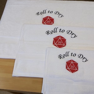 May include: White bath towels featuring the embroidered phrase "Roll to Dry" and a red and white D20 die graphic. The towels are stacked, with the largest at the top and the smallest at the bottom. The towels are made of a soft, absorbent material.