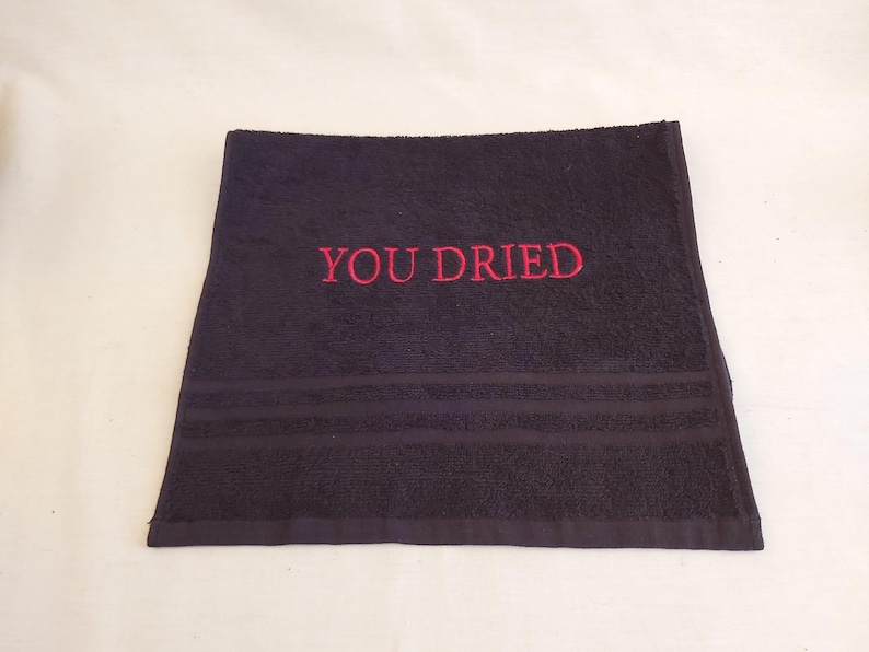Pu&ograve; includere: Un asciugamano grigio scuro con la scritta ricamata in rosso "YOU DRIED". L'asciugamano ha tre strisce orizzontali vicino al bordo inferiore. &Egrave; piegato e appoggiato su una superficie bianca.