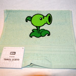 Puede incluir: Una toalla de invitados verde claro con una imagen bordada de una planta verde con boca y ojos. La toalla tiene el texto "GUEST TOWEL" y "Plus" en la etiqueta.