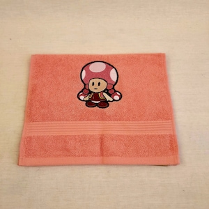 May include: A pink hand towel with an embroidered image of a cartoon mushroom character wearing a red dress.