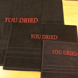 Pu&ograve; includere: Un set di tre asciugamani da bagno neri con la scritta ricamata "YOU DRIED" in rosso. Gli asciugamani sono di diverse dimensioni, il pi&ugrave; grande in alto. Ogni asciugamano ha una superficie strutturata.