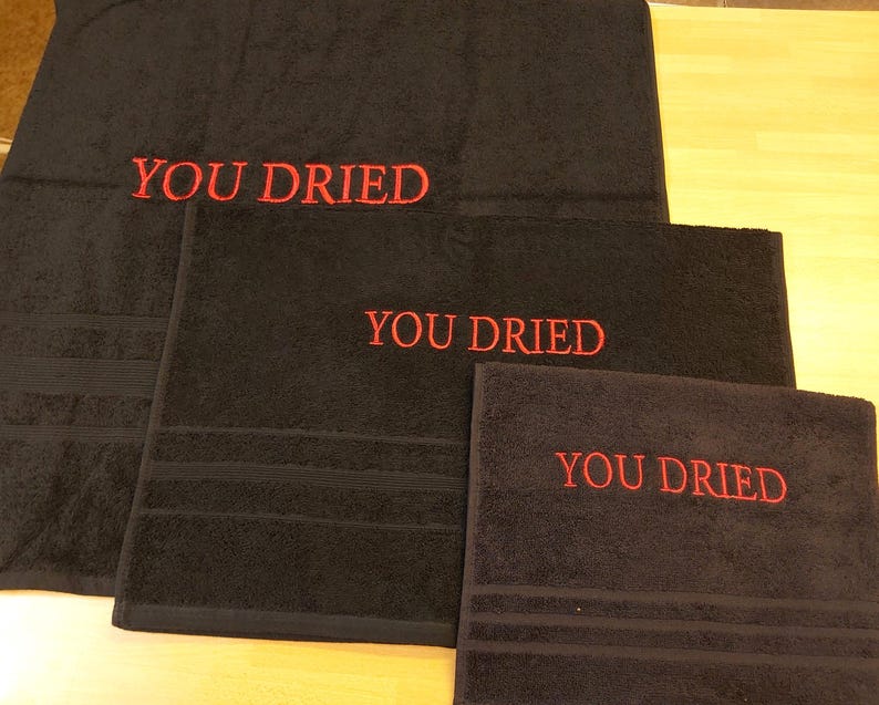 Pu&ograve; includere: Un set di tre asciugamani da bagno neri con la scritta ricamata "YOU DRIED" in rosso. Gli asciugamani sono di diverse dimensioni, il pi&ugrave; grande in alto. Ogni asciugamano ha una superficie strutturata.