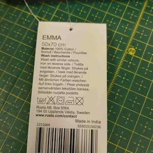 Pu&ograve; includere: Un'etichetta bianca con il nome "EMMA" e dimensioni di 50x70 cm. L'etichetta elenca il materiale come 100% cotone e le istruzioni per il lavaggio. L'etichetta include anche simboli di cura, l'indirizzo del produttore e un codice a barre.