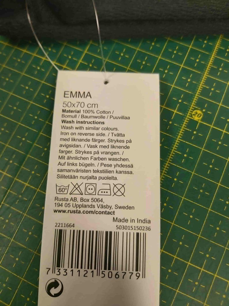 Pu&ograve; includere: Un'etichetta bianca con il nome "EMMA" e dimensioni di 50x70 cm. L'etichetta elenca il materiale come 100% cotone e le istruzioni per il lavaggio. L'etichetta include anche simboli di cura, l'indirizzo del produttore e un codice a barre.