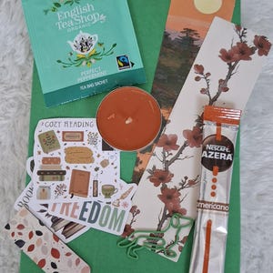 May include: A green-wrapped gift box containing items for a relaxing reading session. It features a peppermint tea bag, a candle, bookmarks, a coffee sachet, and stickers with the words "Cozy Reading" and "Freedom".