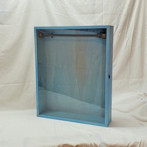 May include: A light blue wooden shadow box with a glass front and a black metal rod inside. The box has a simple design and is perfect for displaying small items.