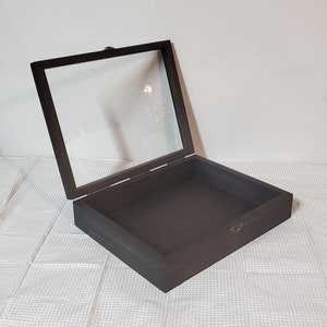 May include: A dark brown, square display box with a hinged glass lid. The box is open, revealing the interior. The lid is attached with a small metal hinge and clasp. The box is sitting on a white patterned surface.