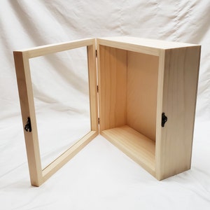 May include: A wooden display case with a glass door. The case is open and the door is hinged on the right side. The case has a small shelf inside. The case is made of unfinished wood and has two small black metal latches.