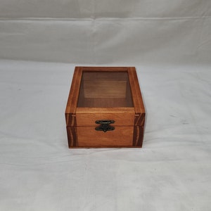 May include: A wooden box with a clear glass top and a metal latch. The box is stained a light brown color and has a wood grain pattern.