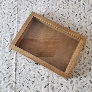 May include: A wooden shadow box with a clear glass insert. The box is light brown and has a simple design.