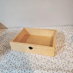 May include: A natural wood box with a hinged lid and a black metal latch. The box is empty and sitting on a white fabric with a black leaf pattern.