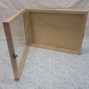 May include: A wooden display case with a glass door and a hinged lid. The case is made of light wood and has a simple design. The case is open and the interior is empty.
