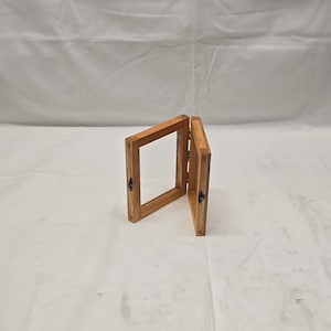 May include: A wooden picture frame with a hinged front, allowing it to open and close. The frame is made of light brown wood and has a simple design.