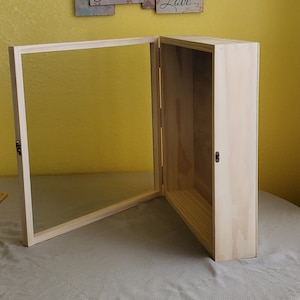 May include: A wooden display case with a glass door, open to reveal an empty interior. The case is made of light wood and has a simple design.