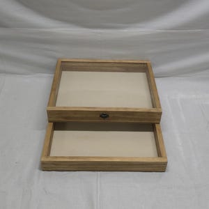 Puede incluir: Una caja de exhibición de madera de dos niveles con una tapa con bisagras y un pequeño pomo de metal. La caja tiene un acabado de madera marrón claro y está vacía.