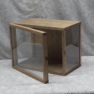 May include: A wooden display cabinet with a glass door. The cabinet is rectangular and has a light brown finish. The door is open, revealing the interior. The cabinet is designed to showcase collectibles or other items.