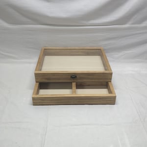 Puede incluir: Una caja de exhibición de madera con tapa de cristal y un cajón. La caja es de madera de color claro y tiene un interior beige. El cajón está dividido en dos compartimentos y asegurado con un cierre metálico.