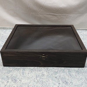 May include: A rectangular dark brown wooden display box featuring a clear glass lid and a metallic latch closure on the front. The box has a visible wood grain texture and a flat top surface, providing a secure container for storage and display.