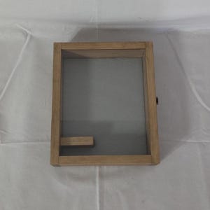 May include: A wooden shadow box with a glass front and a small wooden shelf inside. The box is light brown in color and has a simple design.