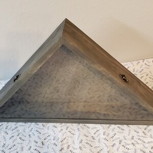 May include: A gray wooden triangular shadow box with a glass front and two brass hinges. The box is sitting on a white fabric with a leaf pattern.