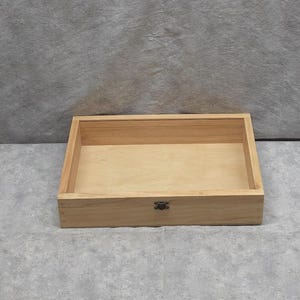 May include: A rectangular wooden box with a light brown finish. The box has a hinged lid and a metal clasp. The interior of the box is empty. The box is sitting on a gray surface.