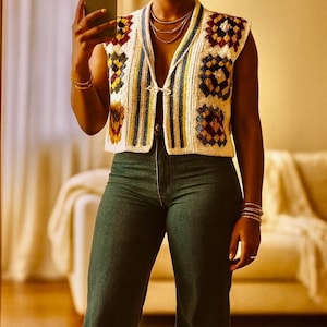 May include: A woman wearing a cream-colored crochet vest with colorful square patterns, paired with dark green flared jeans. The vest has a V-neck and a button closure. She is holding a phone and wearing jewelry.